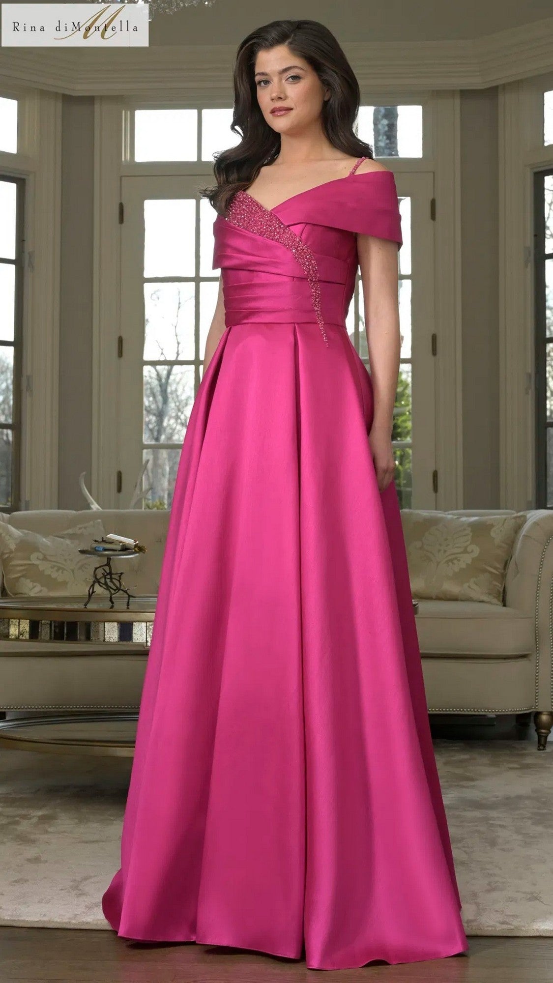 A woman poses indoors by large windows and a beige sofa, wearing the magenta Rina Di Montella RD3221 long formal dress with an off-shoulder design, pleated details, and beaded spaghetti straps.